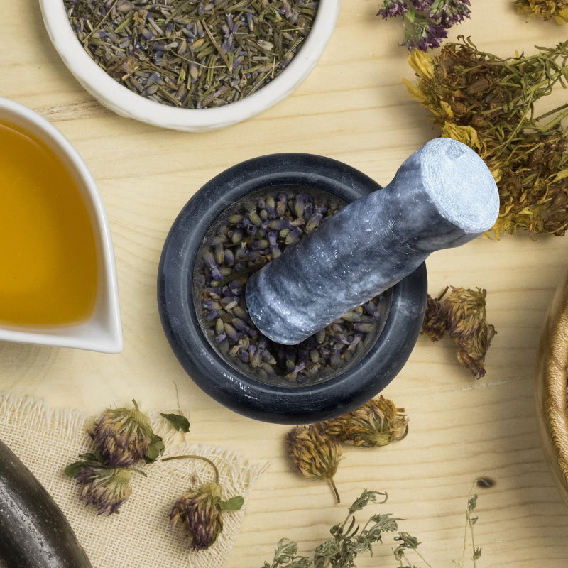 Mortar and pestle with dried herbs on a wooden surface, next to a bowl of tea.